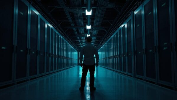 Allée d'une salle des machines déserte, racks d'acier dans une lumière vert-bleu, technicien de dos au loin.