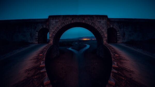 Un vieux pont de pierre se divisant en deux chemins au-dessus d'un lit de rivière asséché, au crépuscule.