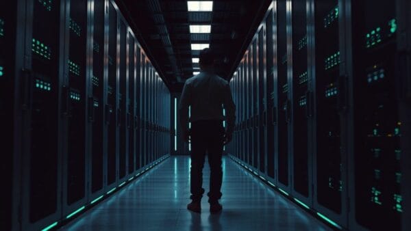 Salle serveurs sombre, racks en acier brossé, silhouette d'un ingénieur de dos au fond de l'allée.