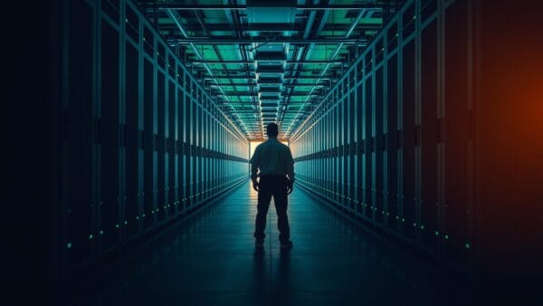 Couloir d'un centre de données avec baies de serveurs et silhouette de technicien de dos au loin.